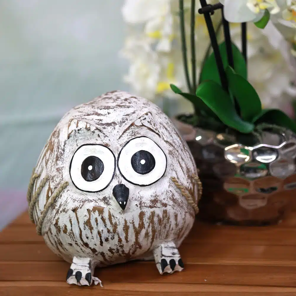 Ein kleines, rundliches Eulen-Dekoobjekt aus weißem und braunem Material mit großen, schwarzen Augen und einem kleinen schwarzen Schnabel steht auf einem Holztisch. Im Hintergrund ist unscharf eine weiße Blume in einem glänzenden, dunklen Topf zu erkennen.