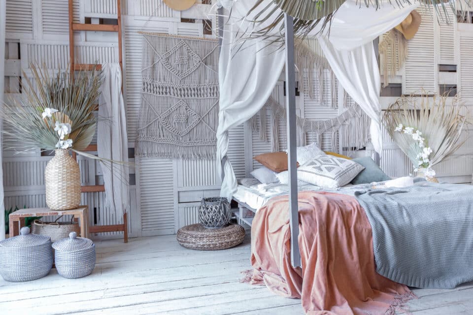 Schlafzimmer im balinesischen Stil, ein Himmelbett mit einer pastellroten und pastellblauen Decke, Kissen und daneben typische geflochtene Gefäße aus Rattan in dezenten Farben.